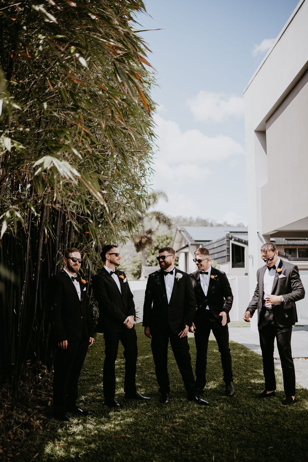 Groomsmen in black tuxedos standing outdoors near bamboo plants on a sunny day.