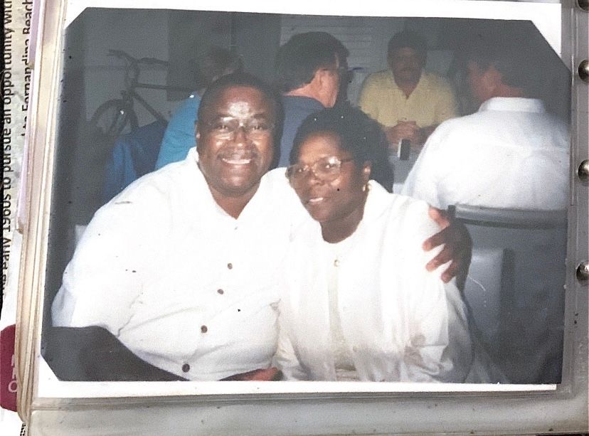 Two people in white clothing sitting together and smiling in what appears to be an indoor setting.