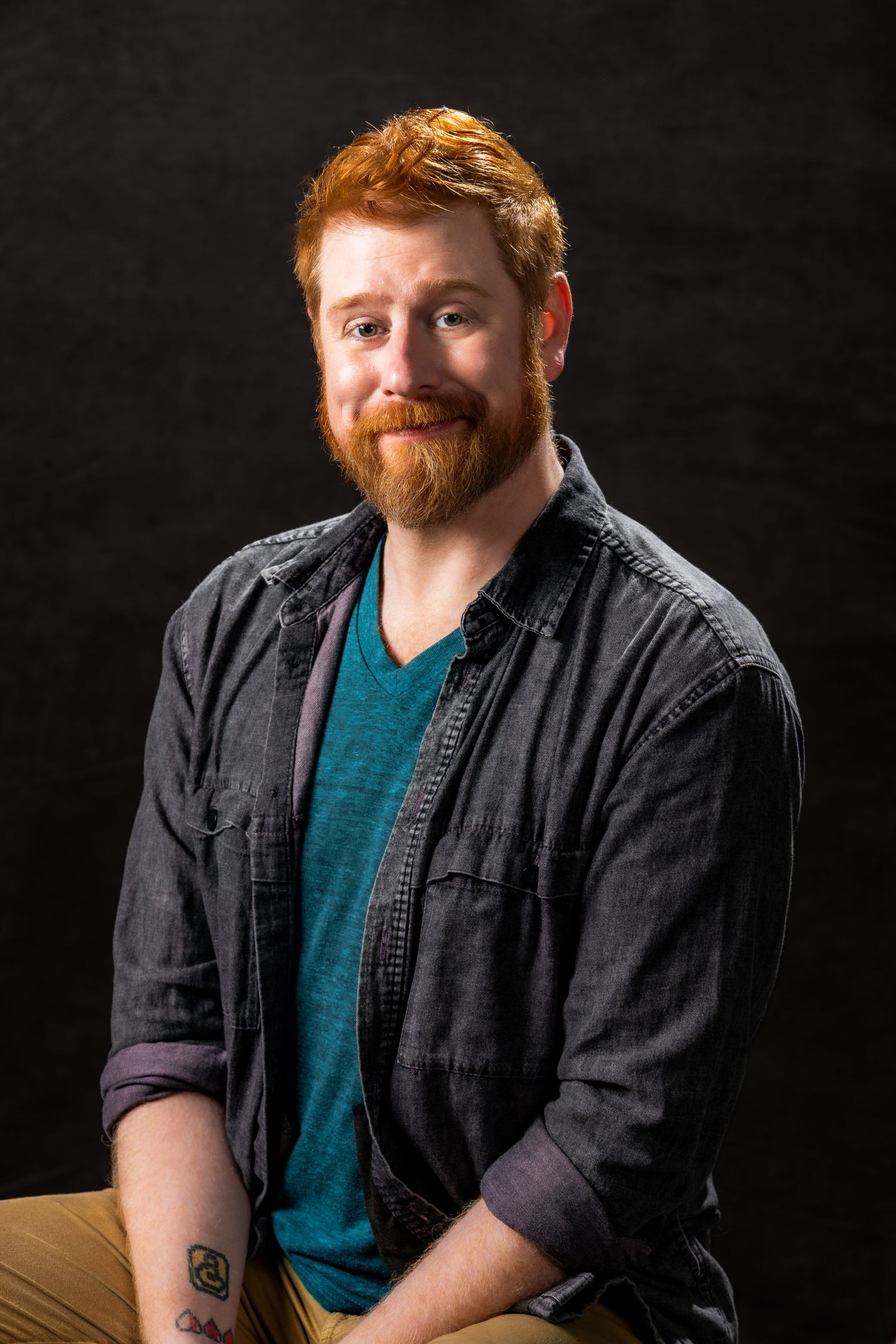 Professional studio portrait showing person in denim button-up shirt and teal t-shirt against dark background.