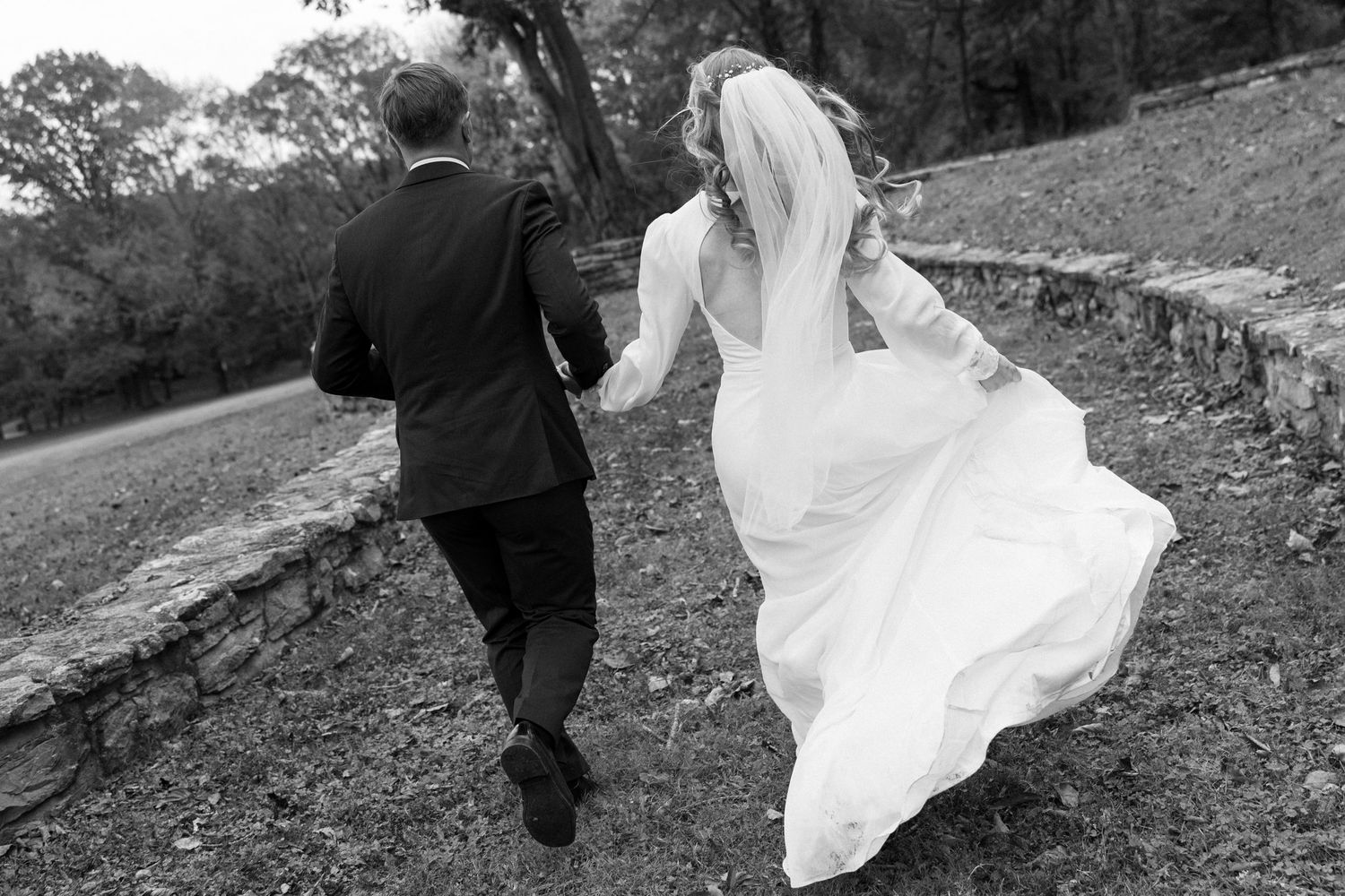A romantic couple in wedding attire runs playfully up a grassy hill during their outdoor photo session.