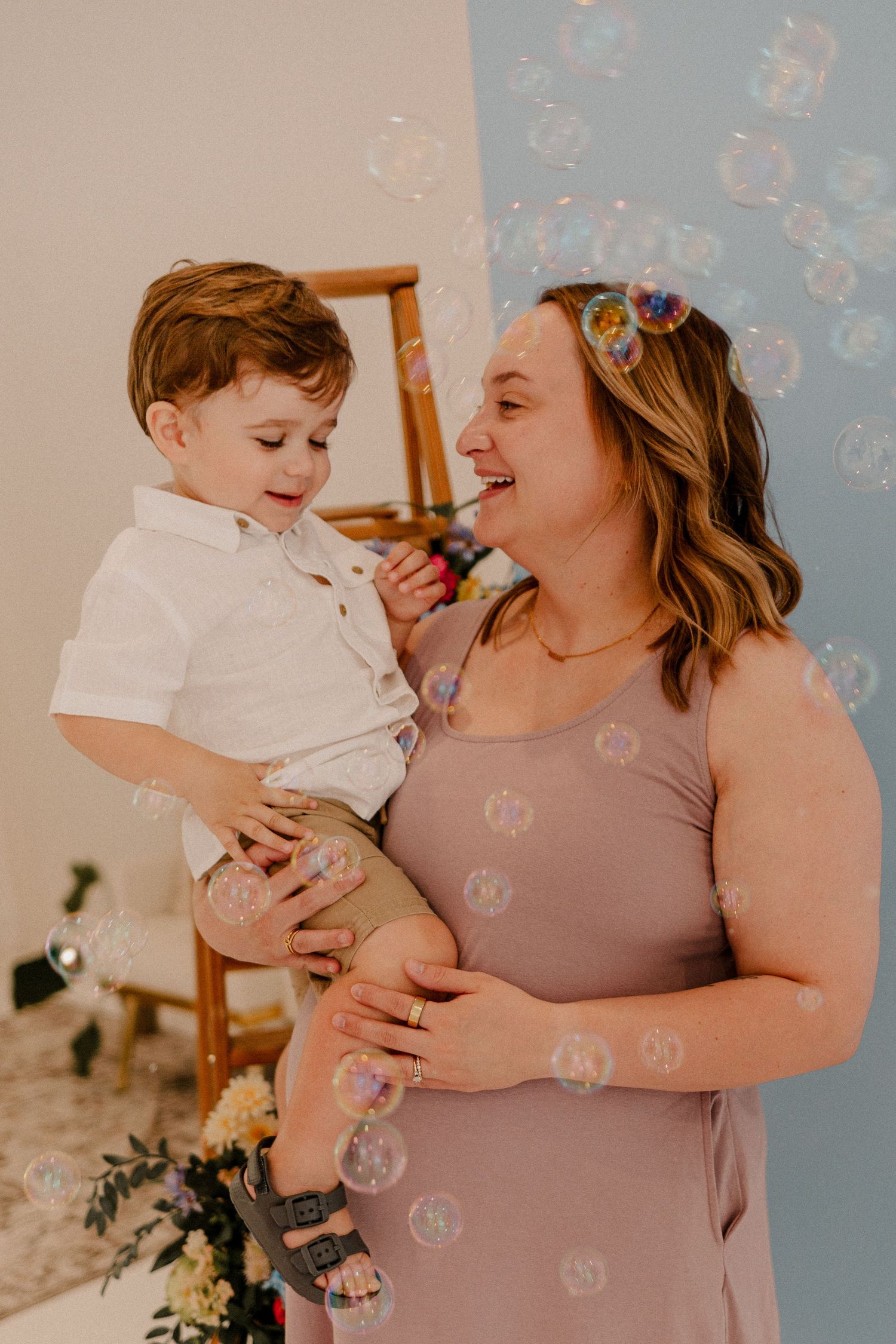 A smiling mother holds her toddler son while colorful bubbles float around them in a bright studio setting.