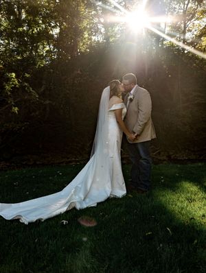 A couple shares a romantic moment in a sunlit garden setting with a flowing white wedding dress trailing behind them.