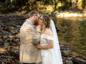 A romantic couple shares a tender kiss in a sunlit forest setting during their outdoor wedding ceremony.