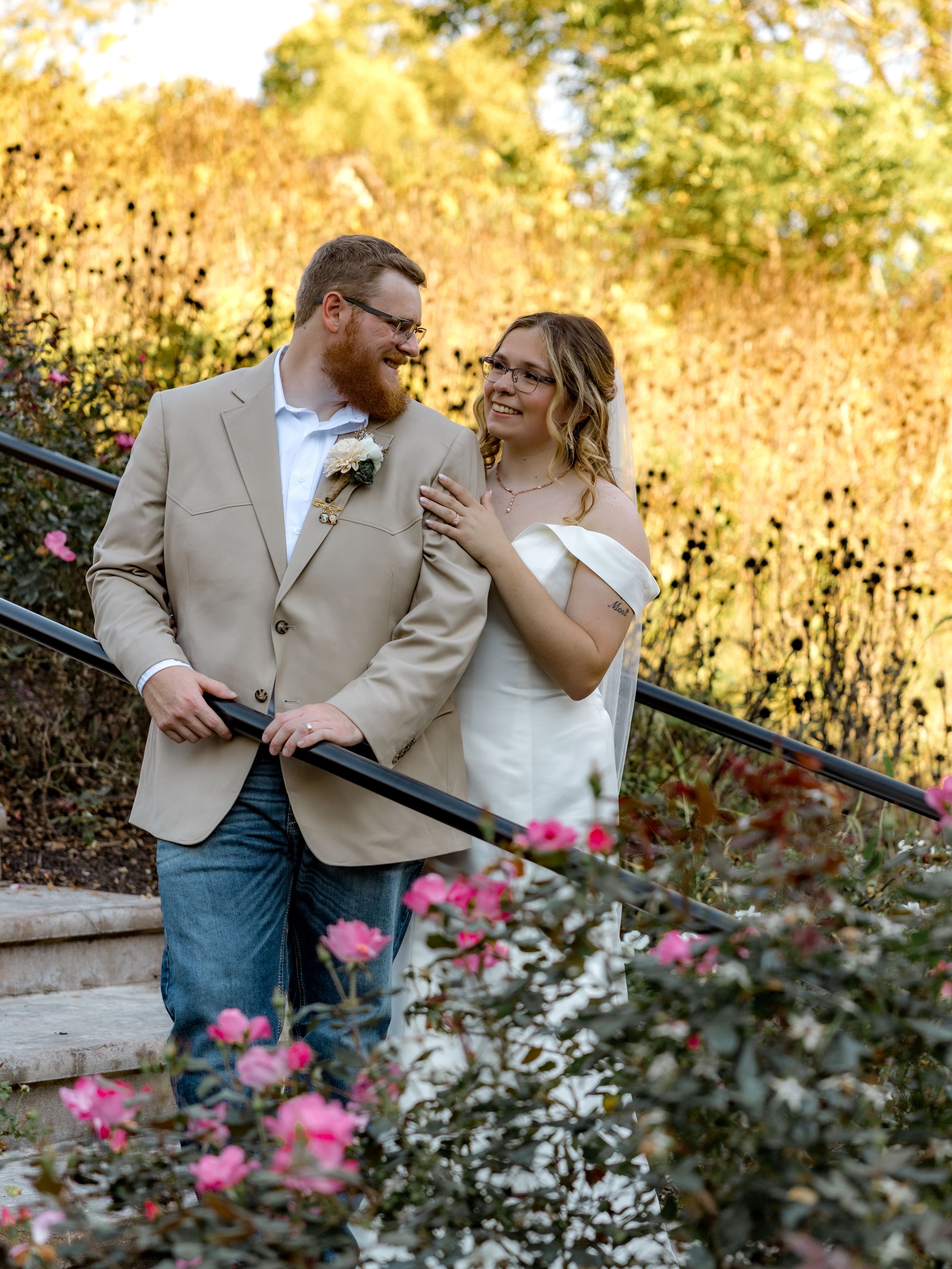 A couple shares a tender moment in a garden with pink flowers and autumn foliage in the background.