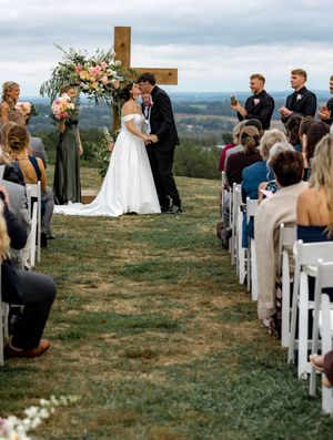 An outdoor wedding ceremony setup on a hilltop with white chairs and a wooden cross altar against a scenic landscape.