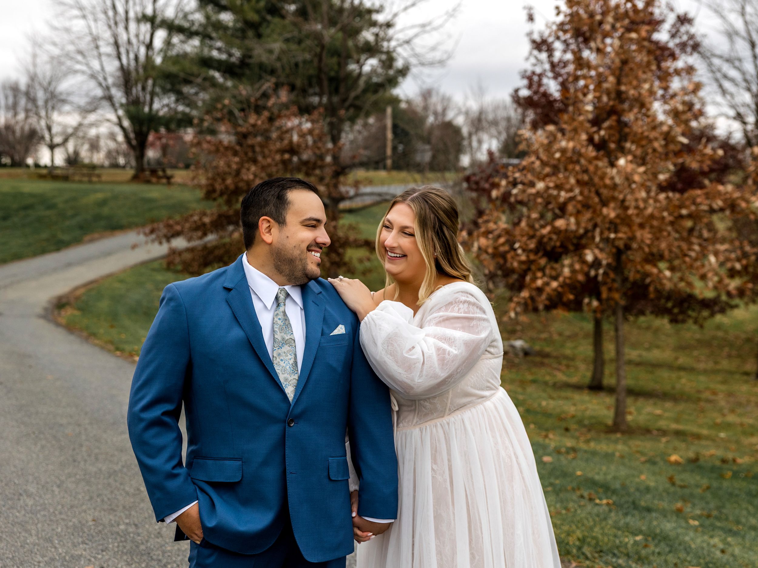 A couple shares a joyful moment on a winding path surrounded by autumn trees in their wedding attire.