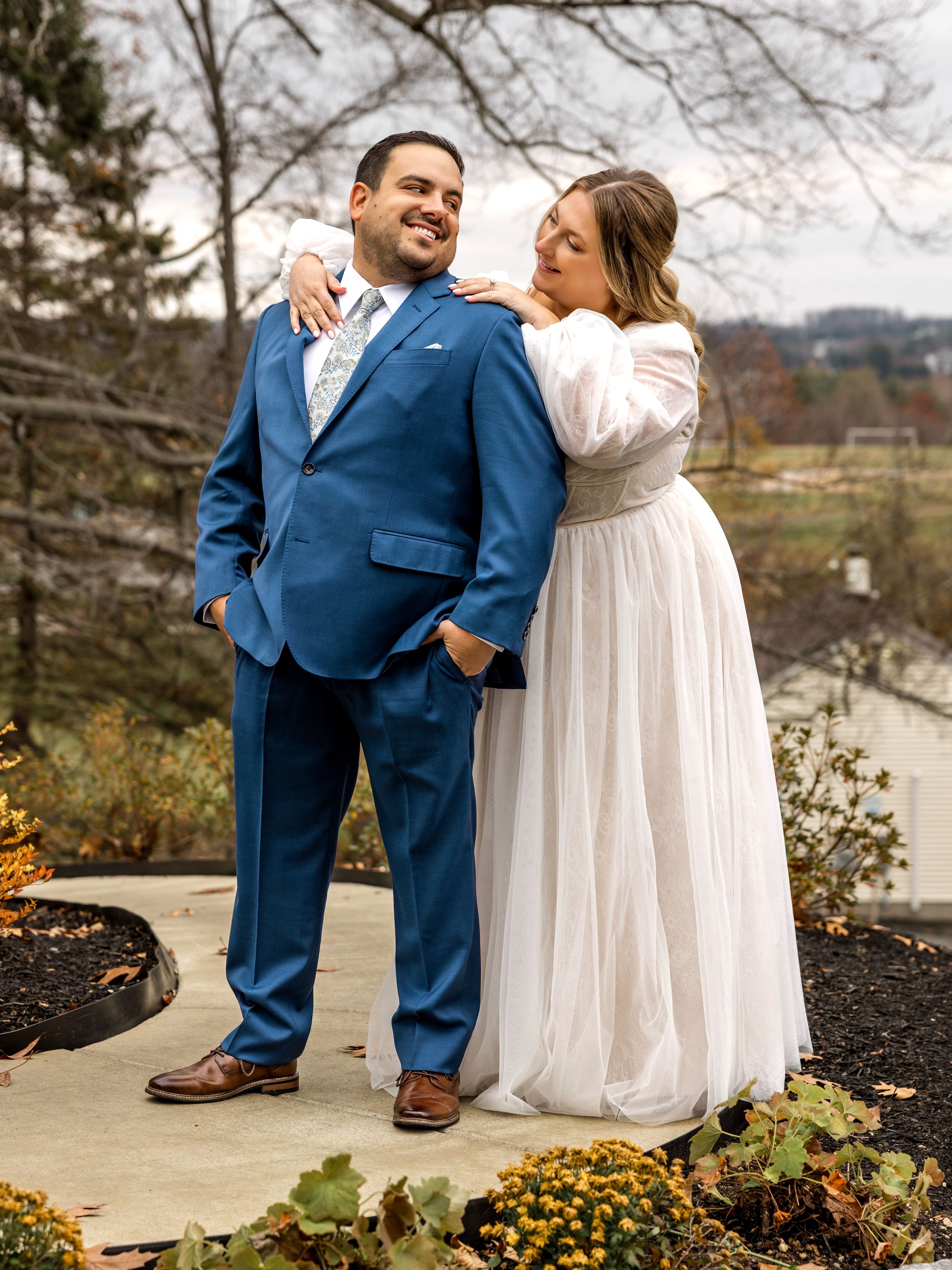 Couple in wedding attire standing close together on a walkway with yellow flowers blooming in the landscaping.
