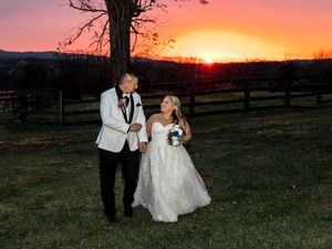 A couple in formal wedding attire walks through a field at sunset while holding their dog.