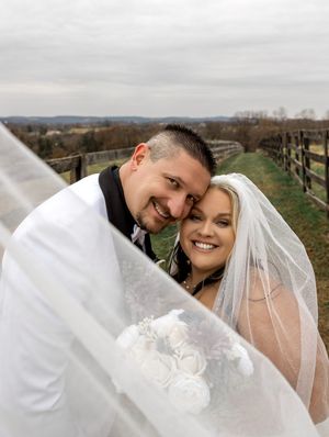A couple in wedding attire share intimate moments under their veil in a rustic outdoor setting with rolling hills.