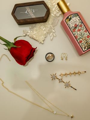 Flat lay of romantic items including a red rose, gold bottle, lace trim, and decorative boxes arranged on a light surface.