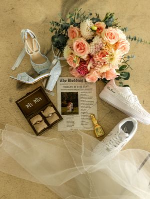 Wedding details flatlay with white shoes, floral bouquet, invitations and delicate tulle veil on beige background.