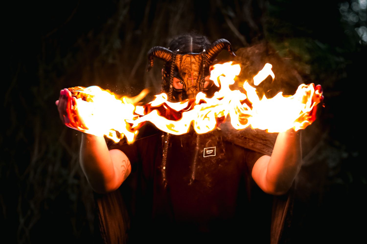 Person in horned demon costume holding flames in both hands in dark outdoor setting.