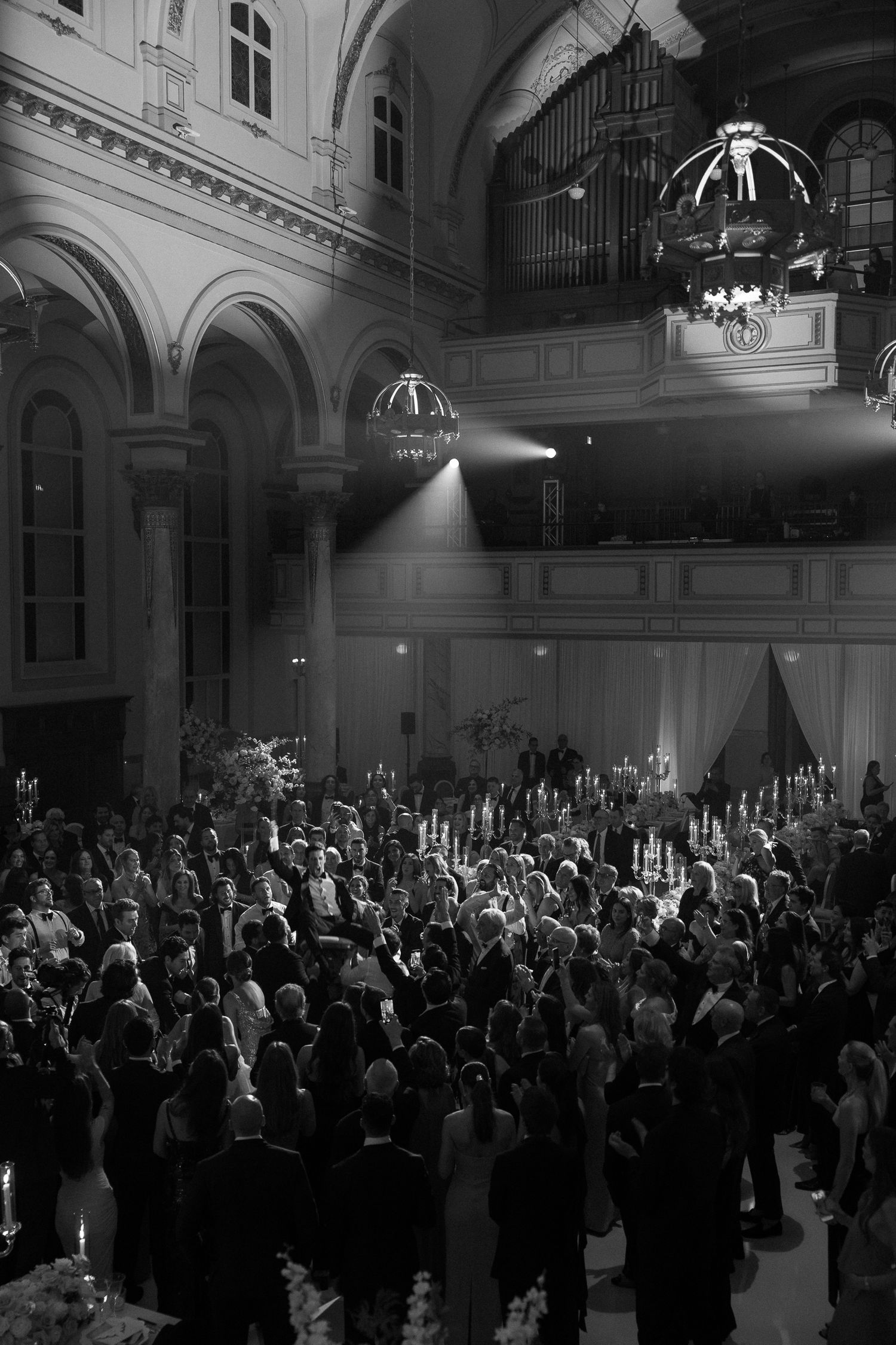 Black and white photo of a grand wedding reception in a historic ballroom with dramatic lighting.