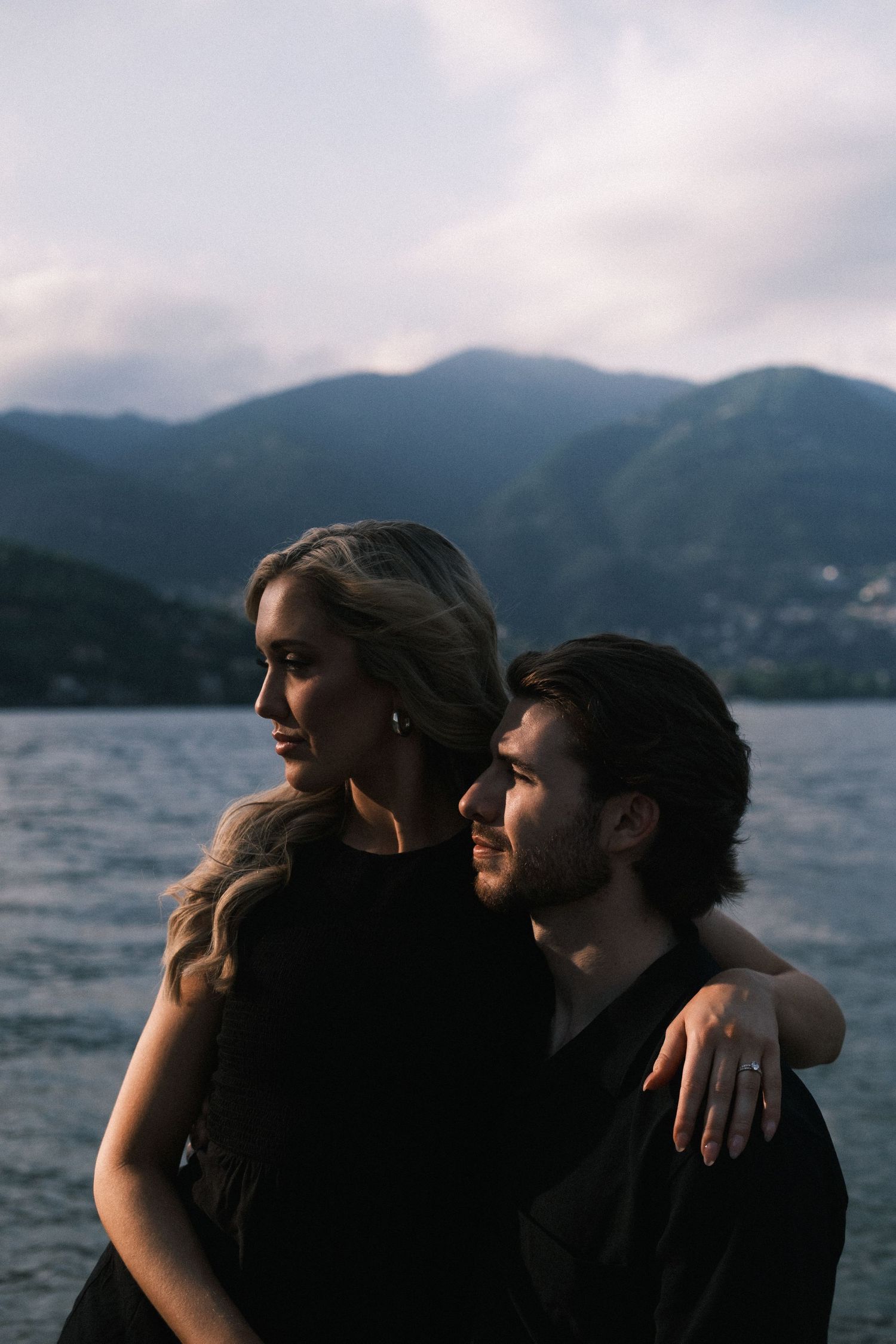 A moody portrait of two figures in dark clothing against a shadowy background, captured in warm dramatic lighting.