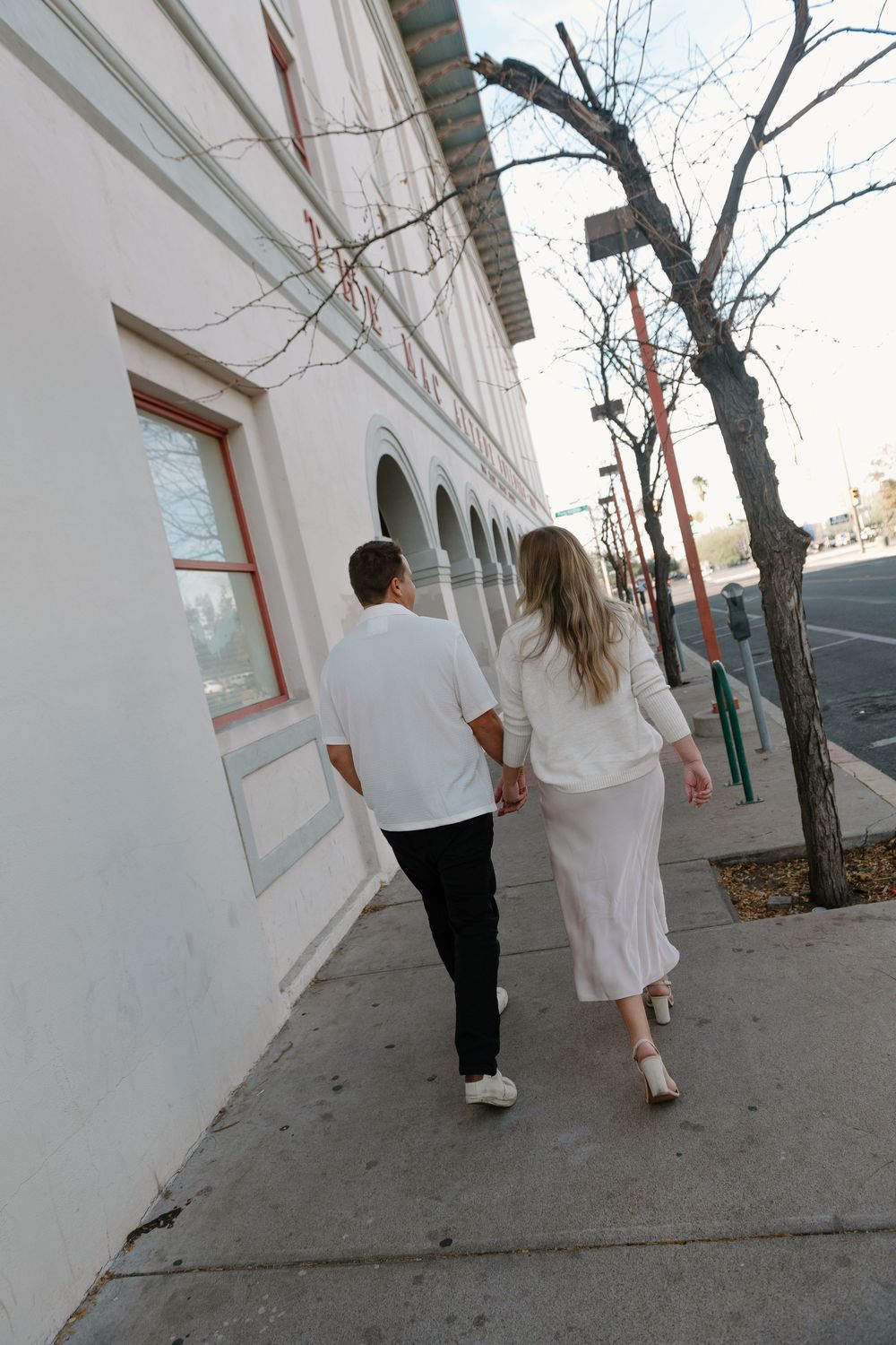 A couple in white clothing walking together along a sidewalk beside a white building wall.