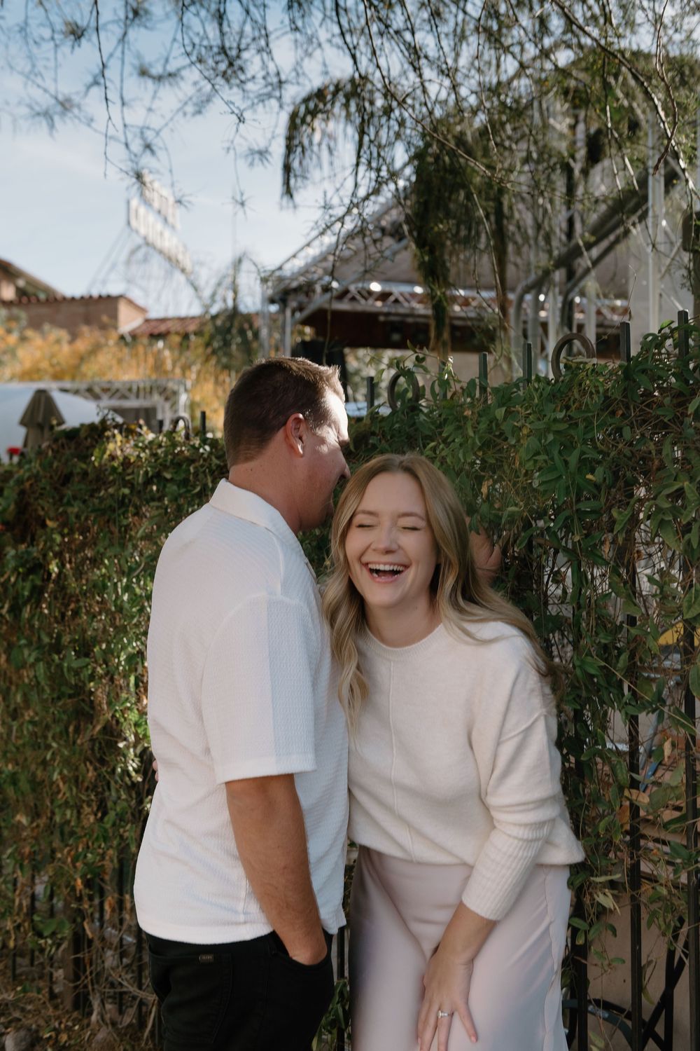 A couple shares a candid moment together while laughing in front of a garden hedge.