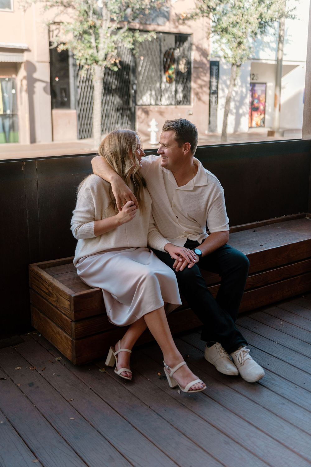 A couple sits intimately together on a wooden bench outside, wearing light colored casual clothing.