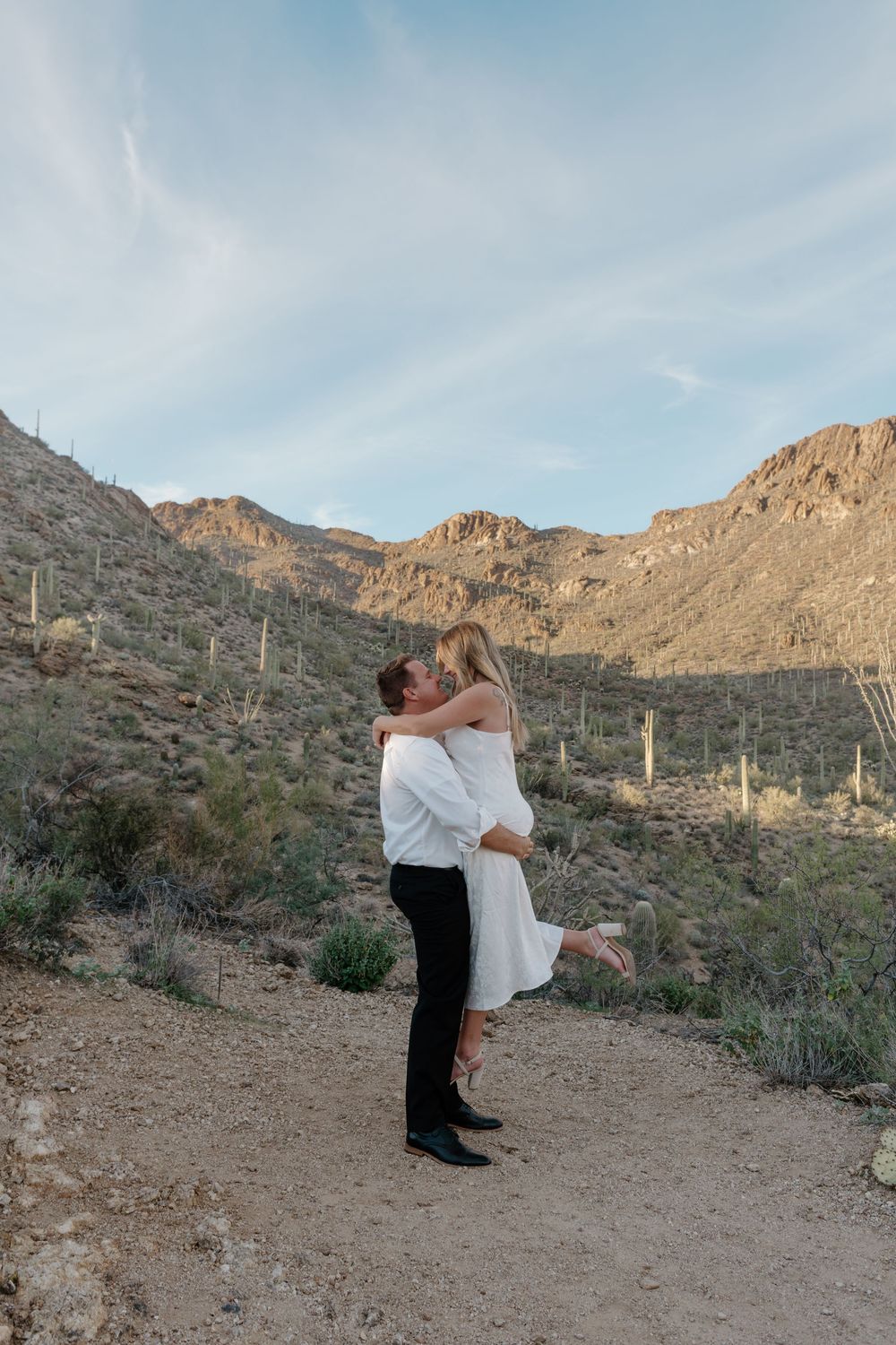 A romantic desert scene with a couple embracing against a backdrop of mountains and saguaro cacti at sunset.