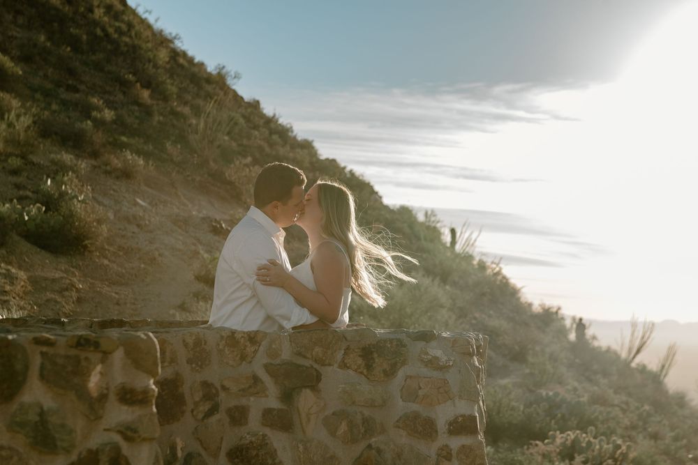 A couple embraces on a rocky coastal cliff during golden hour with sunlight streaming through their hair.
