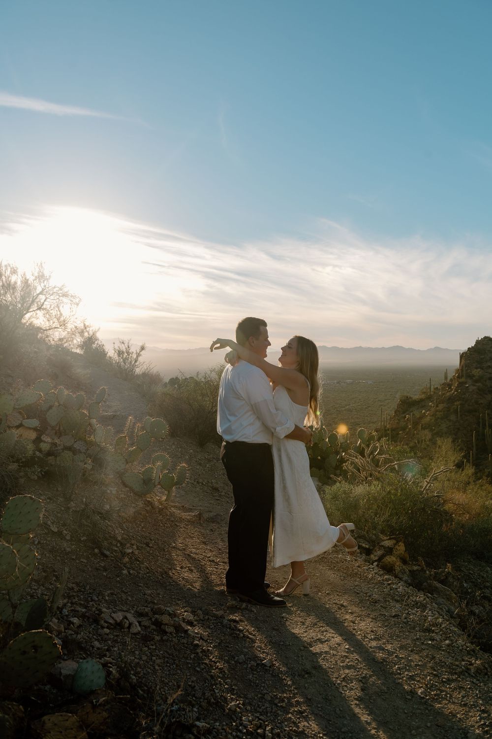 A couple shares a romantic moment at sunset on a desert mountain trail with golden light streaming through the landscape.