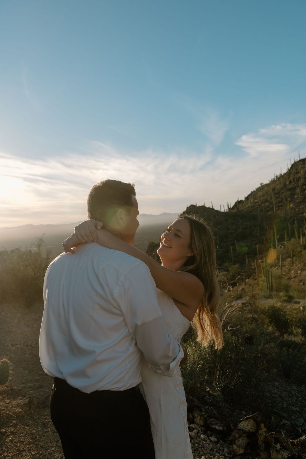 Couple embraces against desert mountain backdrop with saguaro cacti at sunset in warm golden light.