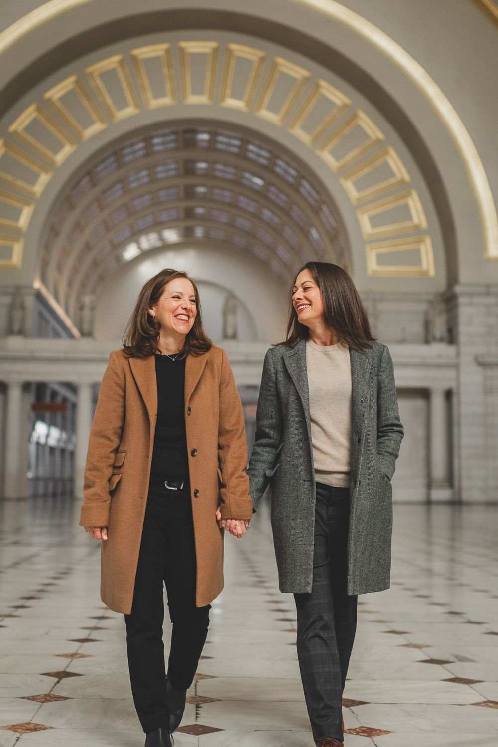 Two newly engagement people in winter coats hold hands and laugh while walking through an elegant hallway of the Union Station Dc