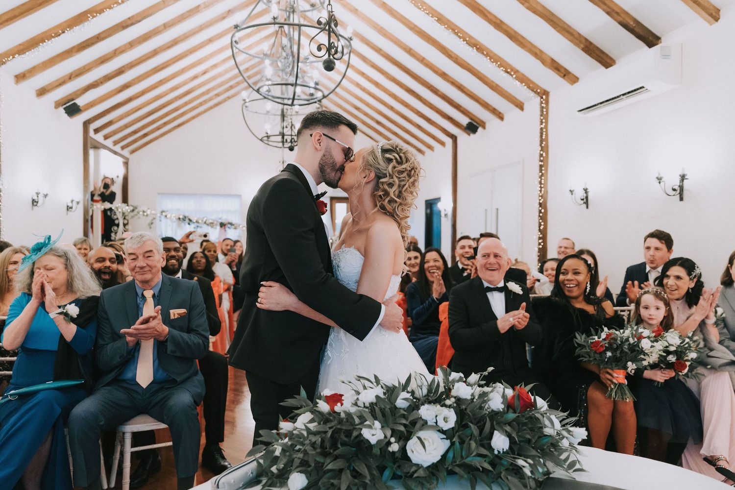 Wedding guests dance and celebrate during a reception under a white tent with exposed wooden beams.