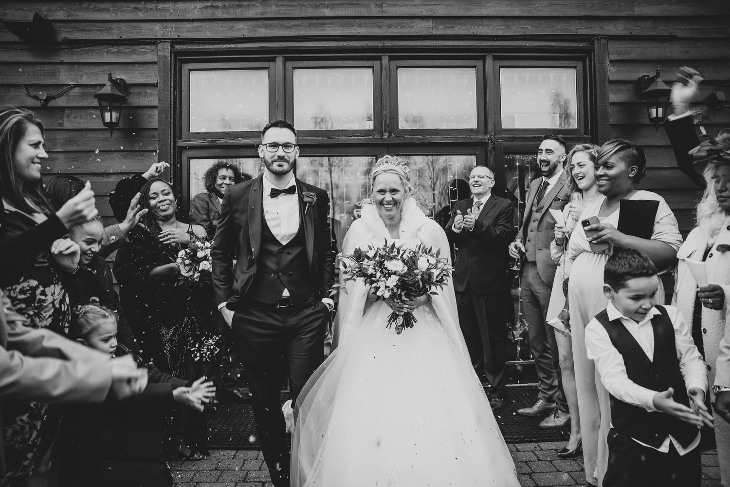 Bride and groom walk through confetti shower surrounded by smiling wedding guests outside the rustic Crondon Park wedding venue.