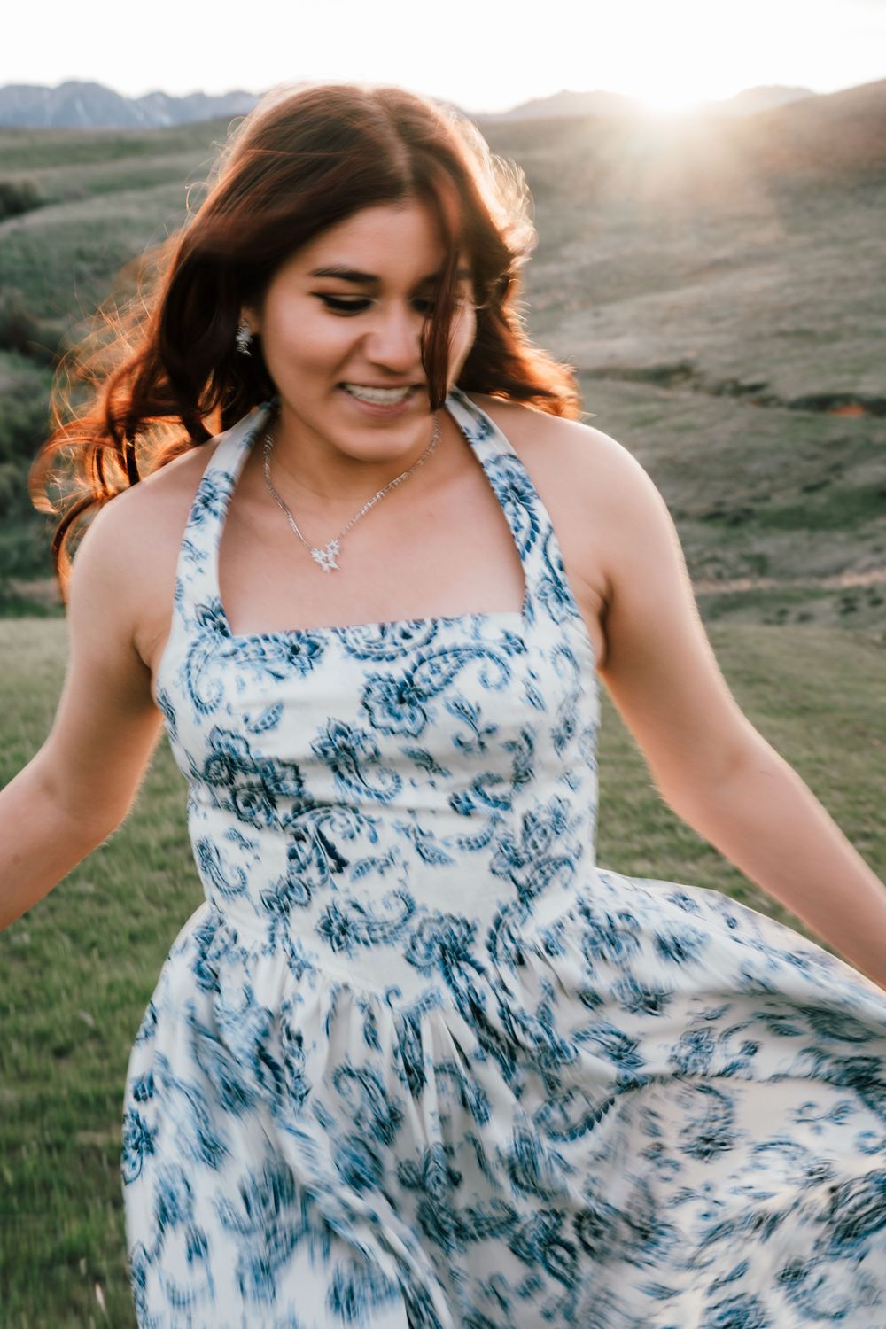 Young woman in blue floral halter dress twirling outdoors at golden hour with scenic hills in background.