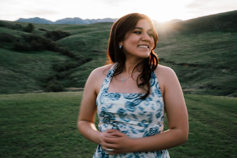 Young woman in floral dress smiling outdoors in green rolling hills at sunset.