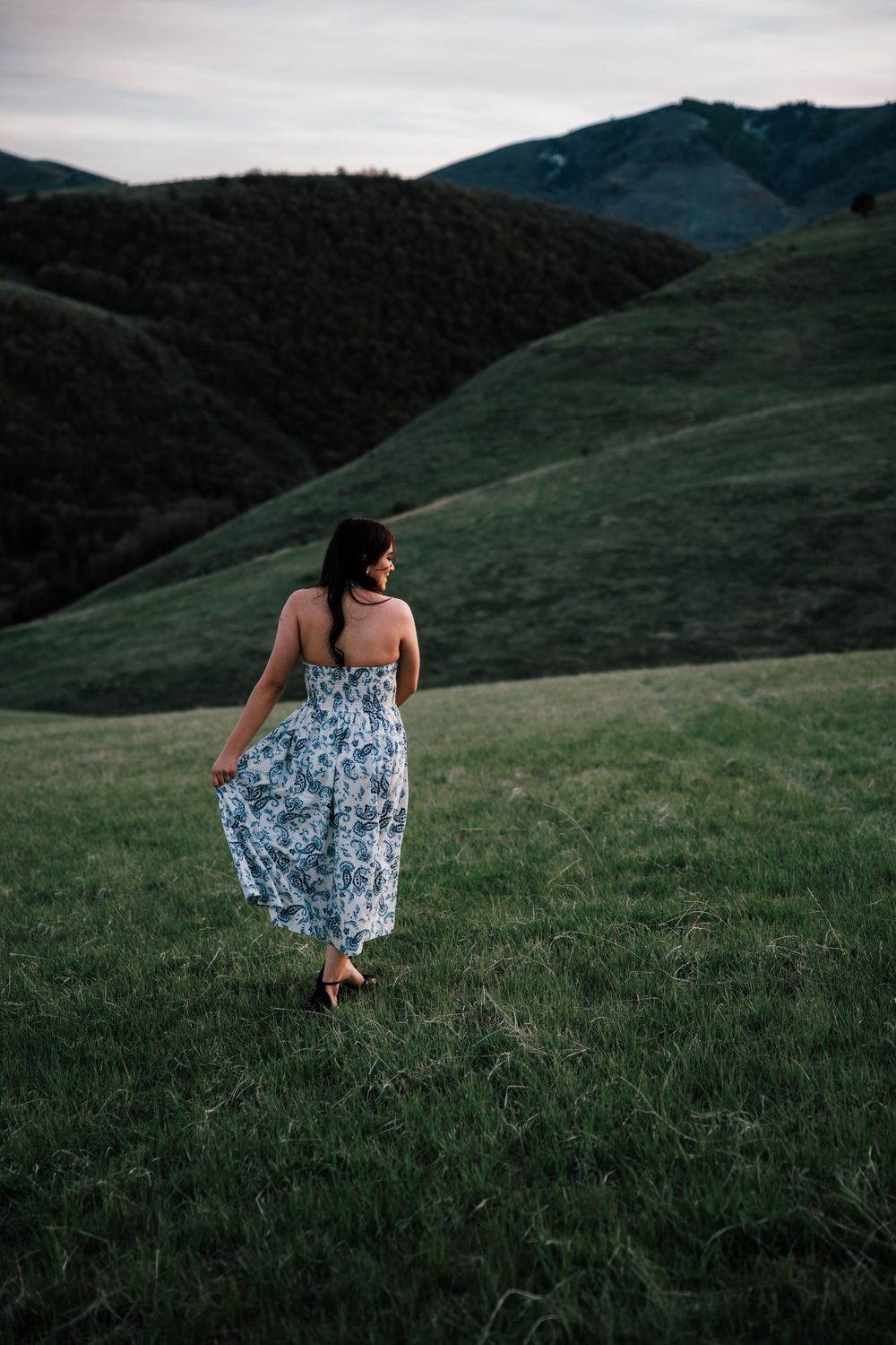 Woman in floral sundress walking through green rolling hills at dusk.