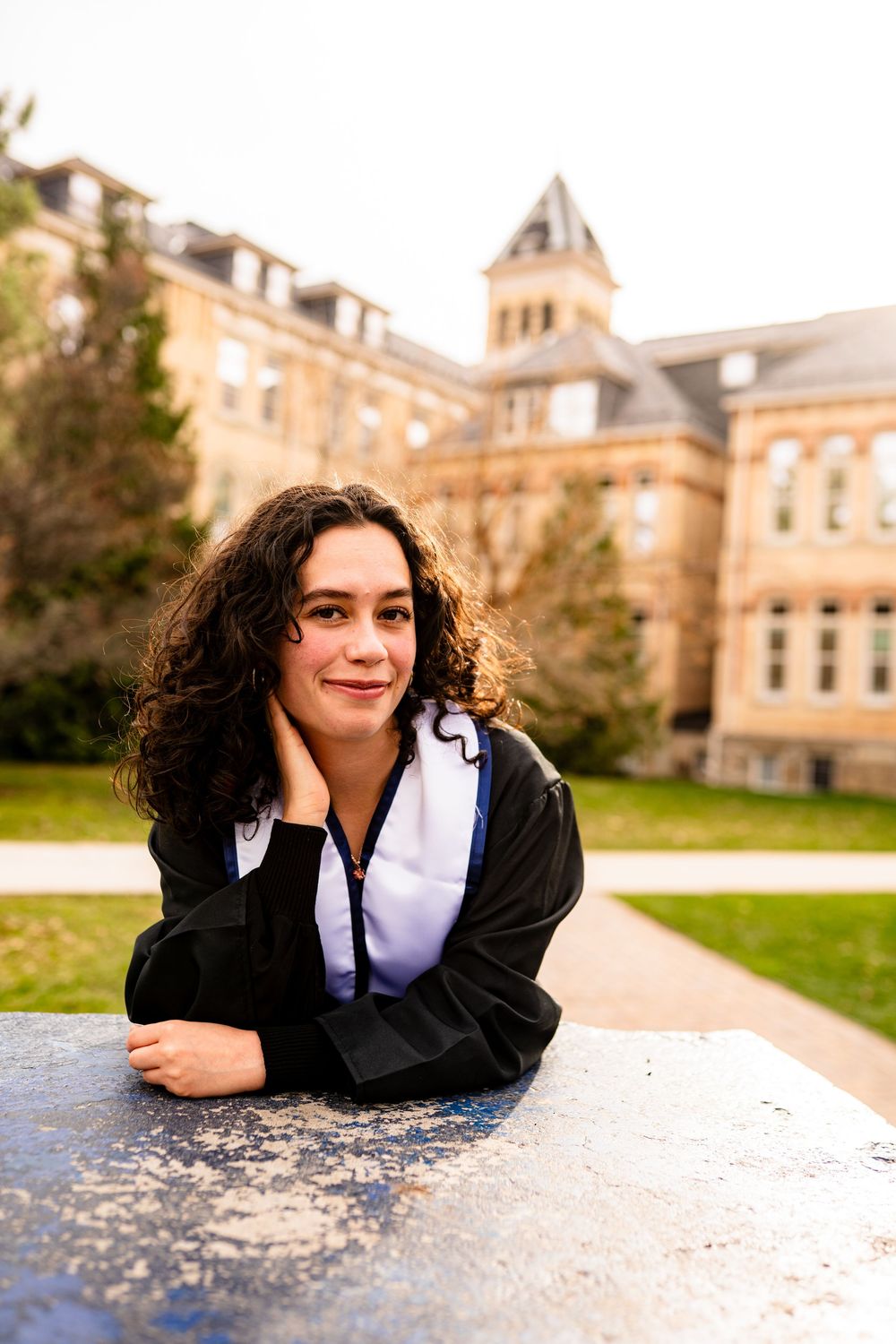 Student in casual attire leaning on table outdoors with historic university building in background.