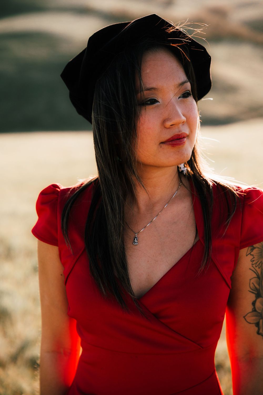 Woman in red wrap dress and black beret with tattoo poses outdoors in golden field at sunset.