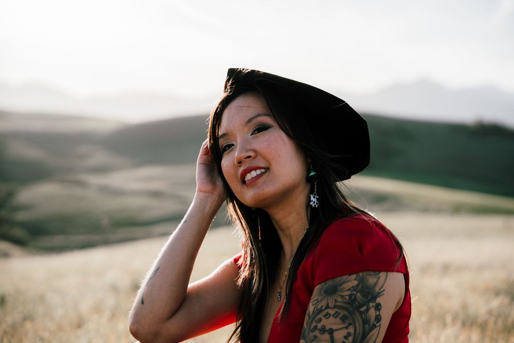 Smiling Asian woman in red top and black beret holds hat in golden field with mountain backdrop.