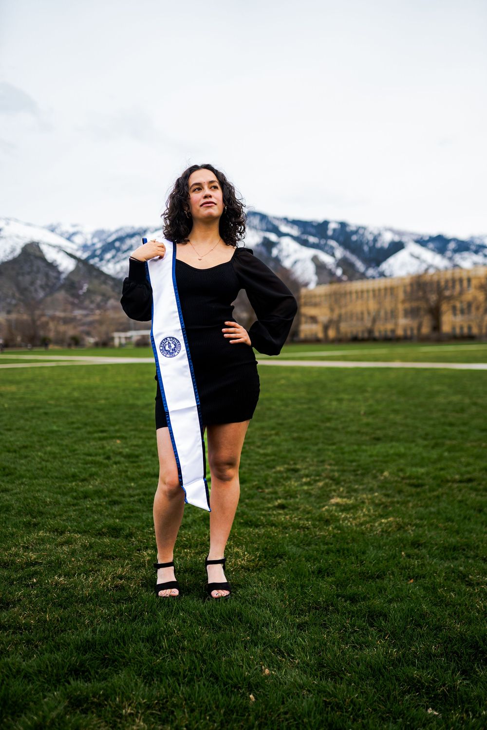 A person in a black dress poses with a snowboard on a grassy field with snow-capped mountains in the background.