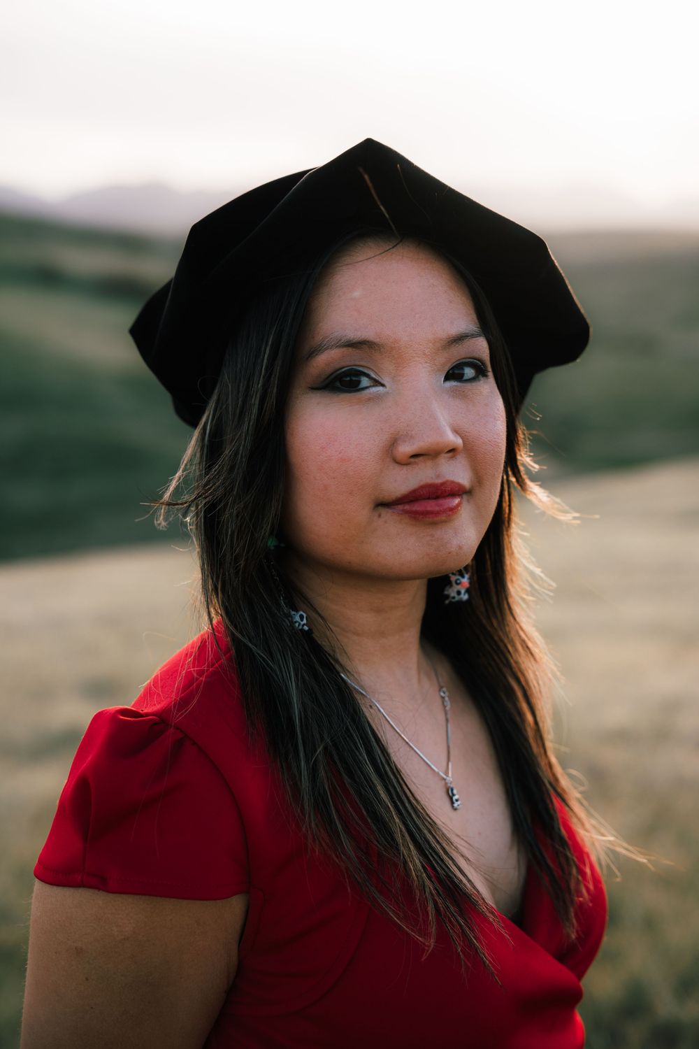 Young Asian woman in red dress and black beret poses outdoors in golden field at sunset.