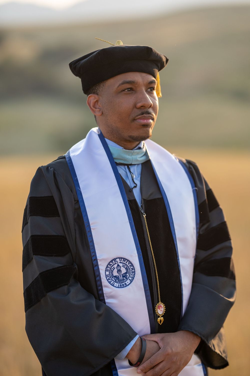 Graduate wearing black academic regalia and white stole stands outdoors during golden hour commencement ceremony.
