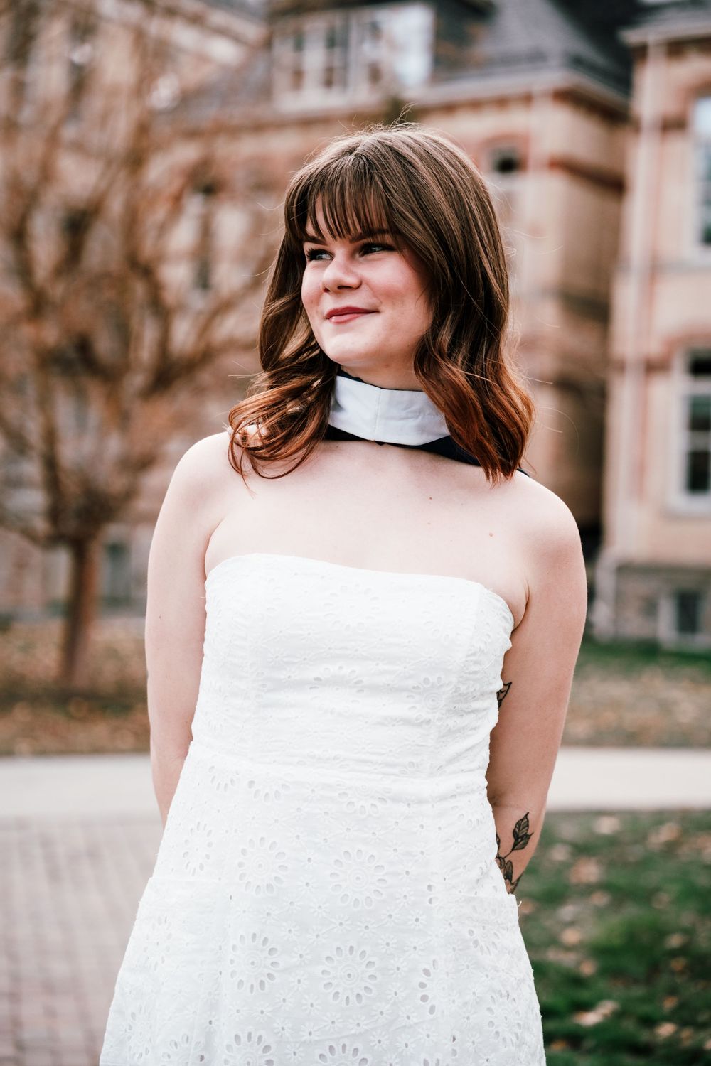 A person in a white lace dress poses outdoors in front of a historic brick building on a fall day.