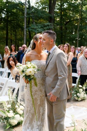 Outdoor wedding ceremony setup with white chairs and floral arrangements under a canopy of trees.