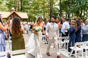 Wedding guests gather outdoors for a celebration under trees with white chairs set up for ceremony.