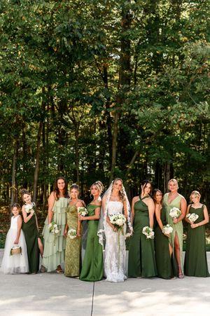 Wedding party in green bridesmaid dresses poses in a line against a wooded backdrop for a panoramic photo.