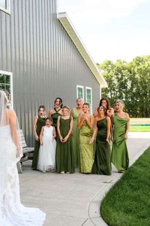 Wedding party in olive green dresses posing together against a modern gray barn exterior.