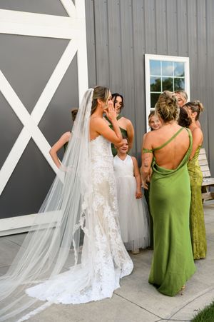 Wedding party in green dresses helps adjust bride's long white veil against gray barn wall in sequence of photos.