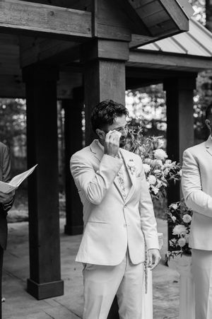 Wedding party members in light colored suits stand together under a covered outdoor area.