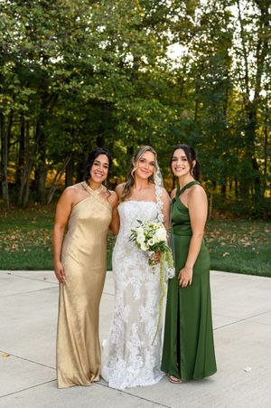 Three people in formal attire pose outdoors in a wooded setting wearing gold, white lace, and emerald green dresses.