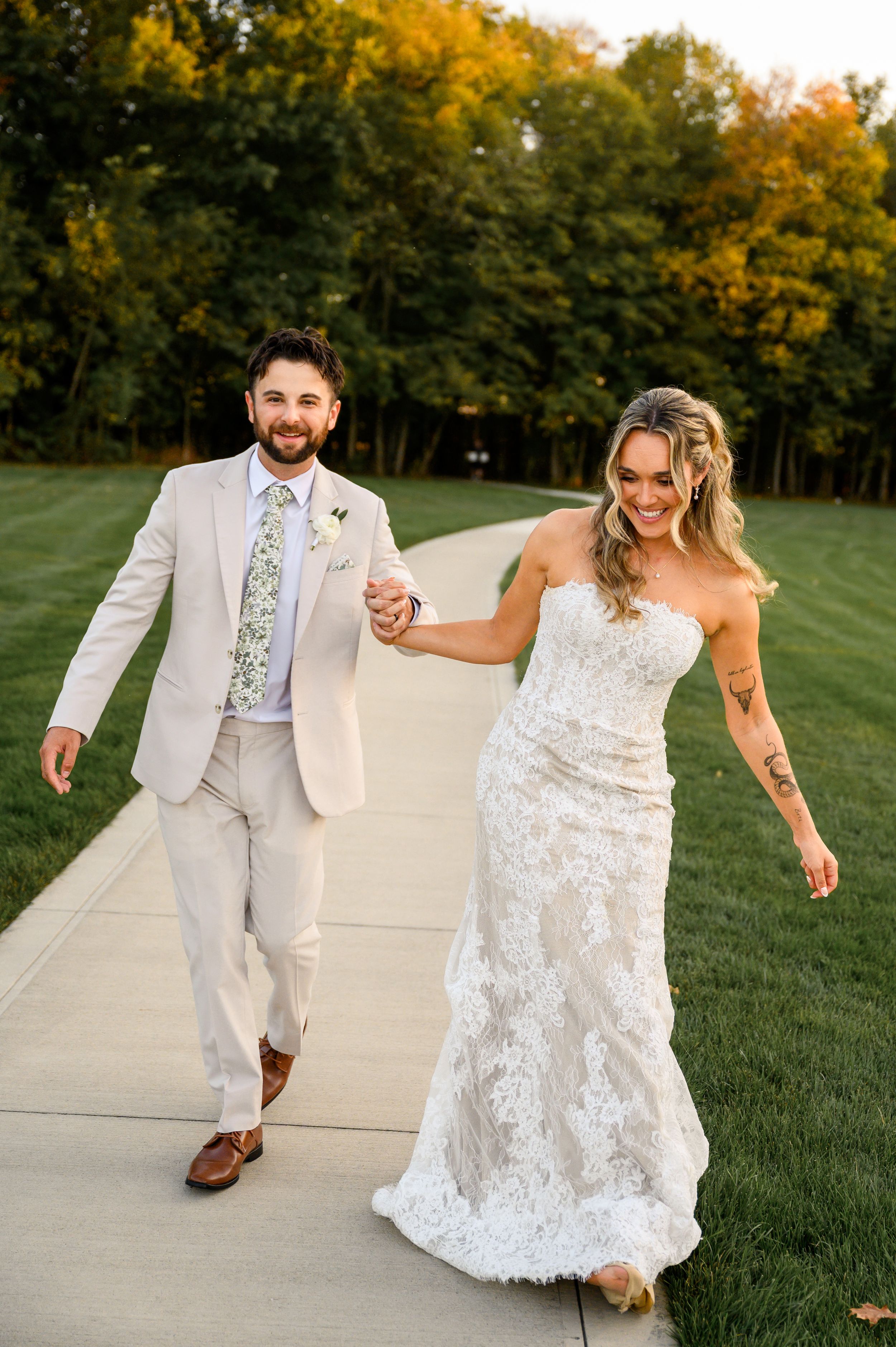 A couple in wedding attire dances playfully on a pathway surrounded by autumn trees and green grass.