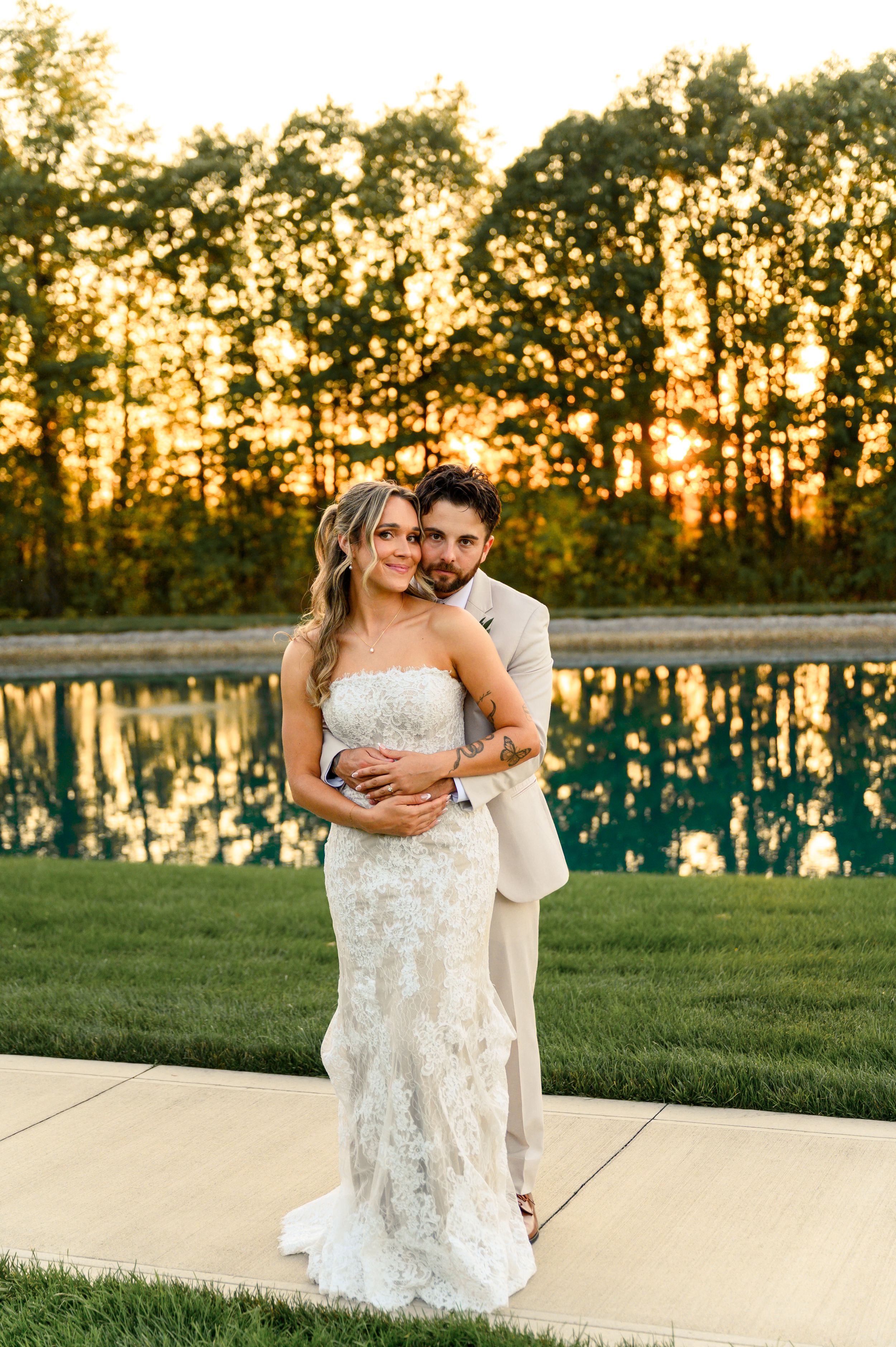 Wedding couple embraces during golden hour sunset by a reflective pool on their special day.