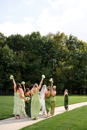 Wedding party in sage green dresses celebrates on a grassy lawn with bouquets raised in the air.