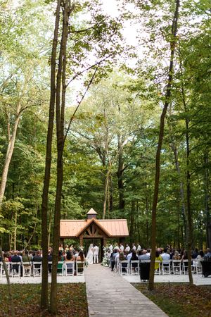 Outdoor forest wedding ceremony setup with white chairs arranged in rows among tall trees.