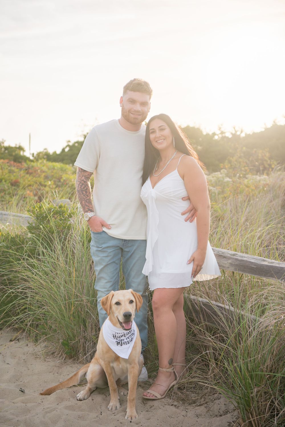 Couple poses with their lab dog on sandy beach trail at dusk.